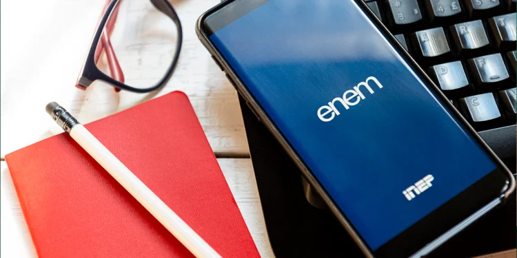 Celular mostrando tela azul do ENEM sobre teclado preto, ao lado de caderno vermelho, lápis e óculos.