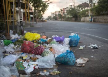 Montanha de sacos de lixo coloridos (azul, vermelho, amarelo) acumulados no calçamento de uma rua urbana vazia ao entardecer.
