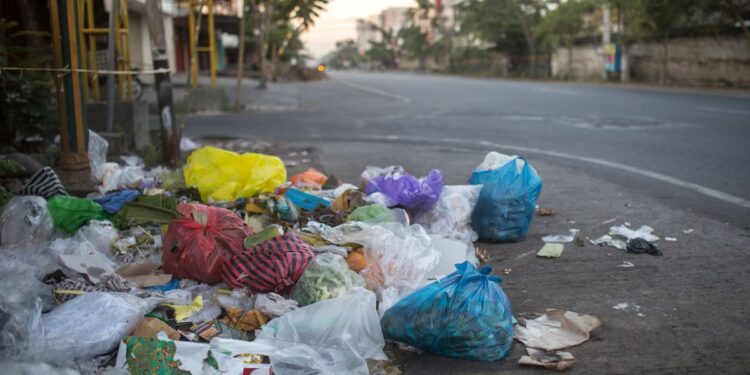 Montanha de sacos de lixo coloridos (azul, vermelho, amarelo) acumulados no calçamento de uma rua urbana vazia ao entardecer.