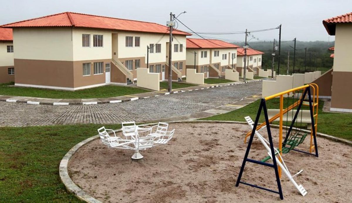 Vista lateral de casas populares geminadas com telhado cerâmico e playground infantil em primeiro plano, contendo um gira-gira branco e um balanço.