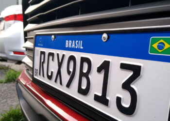 Placa Mercosul com fundo branco, faixa azul com "BRASIL" e bandeira, código RCX9B13 em caracteres pretos.