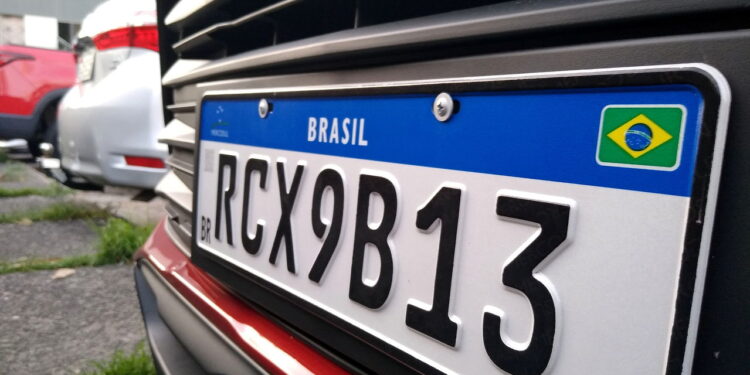 Placa Mercosul com fundo branco, faixa azul com "BRASIL" e bandeira, código RCX9B13 em caracteres pretos.