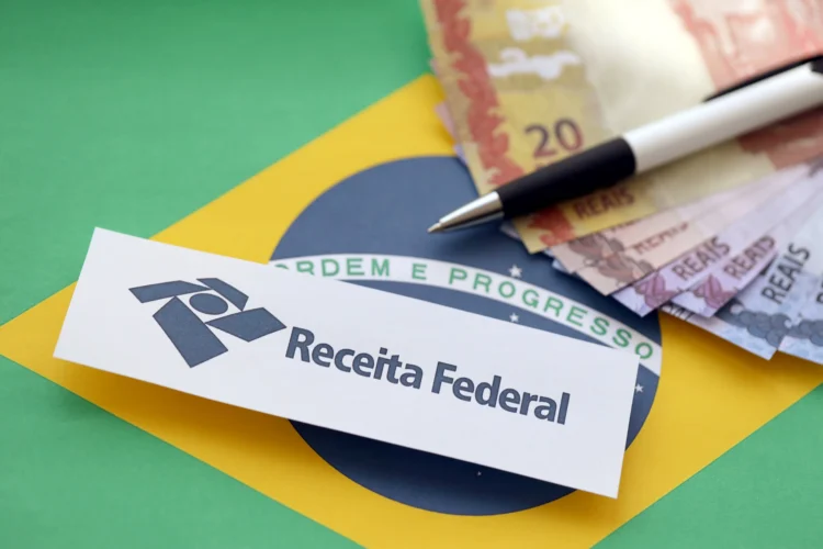 Logo da Receita Federal sobre bandeira do Brasil com notas de real e caneta esferográfica preta.