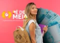 Uma jovem estudante sorridente com mochila bege nas costas e segurando livros roxos. Ao lado, o logotipo colorido do programa Pé-de-Meia com o slogan "A poupança do ensino médio" sobre um fundo rosa. No fundo, notas de 100 reais dispostas em leque.