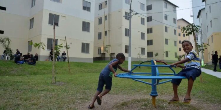 Crianças brincando em um gira-gira no pátio de um conjunto habitacional do Minha Casa, Minha Vida, com prédios de apartamentos ao fundo e gramado.