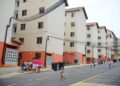 Vista de conjunto habitacional popular com prédios de quatro andares em cores bege e laranja, com moradores e crianças na rua em frente aos prédios.