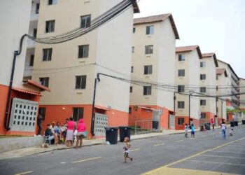 Vista de conjunto habitacional popular com prédios de quatro andares em cores bege e laranja, com moradores e crianças na rua em frente aos prédios.