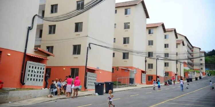 Vista de conjunto habitacional popular com prédios de quatro andares em cores bege e laranja, com moradores e crianças na rua em frente aos prédios.