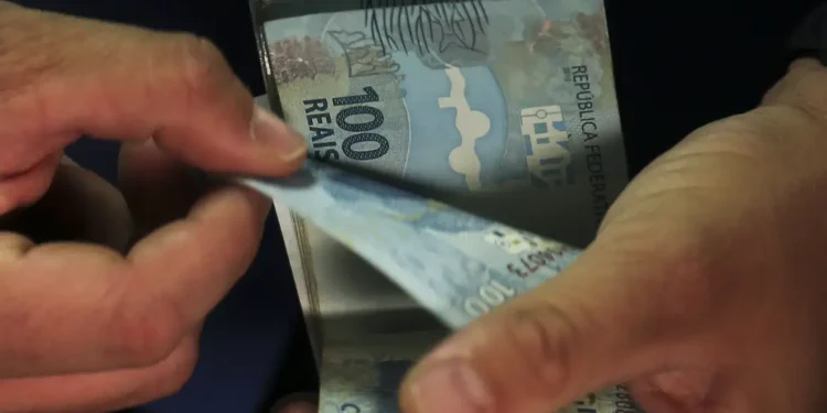 Mãos contam cédulas de R$ 100 do real, representando o pagamento da parcela do Bolsa Família aos beneficiários do programa.