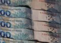 Close em várias notas de 100 reais (cem reais) empilhadas, mostrando o logotipo do Banco Central do Brasil e detalhes da estampa de corais azuis.