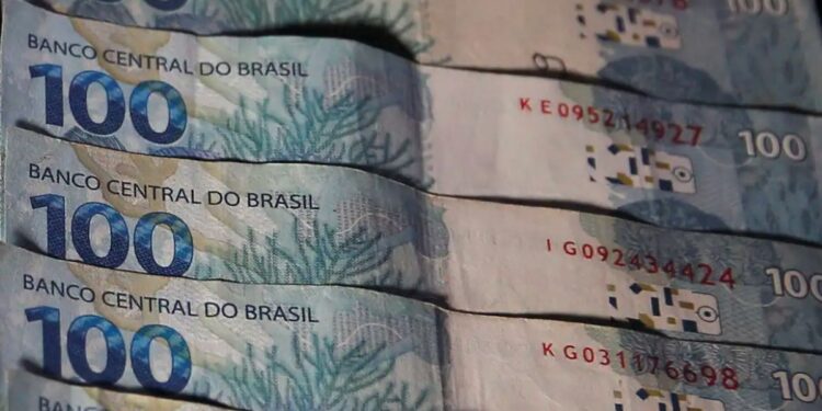Close em várias notas de 100 reais (cem reais) empilhadas, mostrando o logotipo do Banco Central do Brasil e detalhes da estampa de corais azuis.
