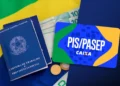 Carteira de trabalho azul, notas de R$ 100, moedas e cartão PIS/Pasep Caixa sobre tecido da bandeira do Brasil.