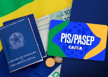 Carteira de trabalho azul, notas de R$ 100, moedas e cartão PIS/Pasep Caixa sobre tecido da bandeira do Brasil.