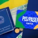 Carteira de trabalho azul, notas de R$ 100, moedas e cartão PIS/Pasep Caixa sobre tecido da bandeira do Brasil.