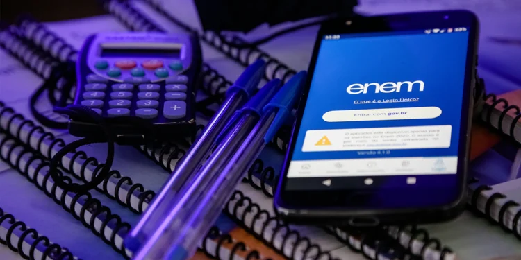 Smartphone exibindo aplicativo do Enem com tela de login gov.br sobre cadernos espirais, canetas azuis e calculadora.