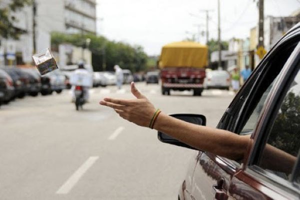 Braço de uma pessoa saindo pela janela de um carro em movimento, jogando uma caixa de papelão vazia na rua. O fundo mostra uma via urbana com outros veículos e pedestres.