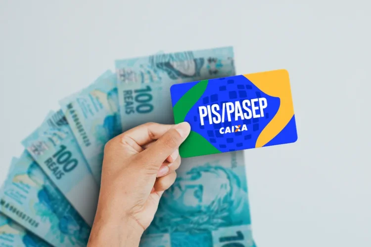 Mão segura cartão PIS/PASEP da Caixa com design verde, azul e amarelo sobre notas de R$ 100.