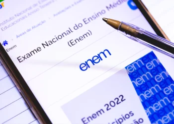 Close de um smartphone exibindo a página oficial do Exame Nacional do Ensino Médio (Enem) no portal gov.br. Sobre a tela, há uma caneta esferográfica de corpo transparente. O fundo é de papel pautado, reforçando o ambiente de estudos.