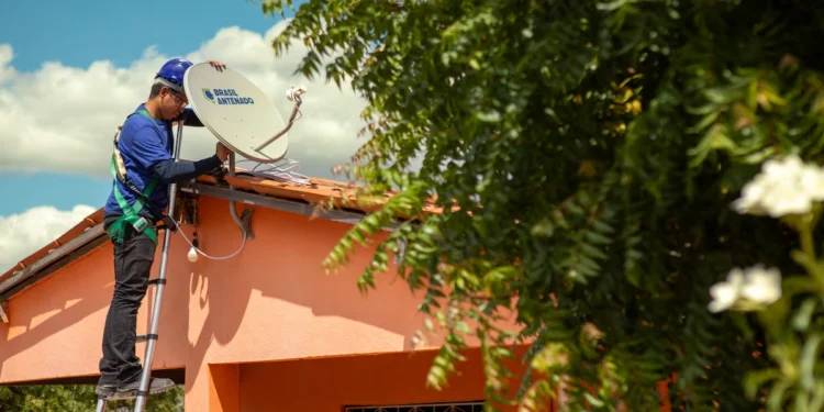 Técnico com capacete azul instala antena parabólica digital do programa Brasil Antenado em telhado de casa laranja.