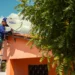 Técnico com capacete azul instala antena parabólica digital do programa Brasil Antenado em telhado de casa laranja.
