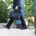 Homem de terno preto e sapatos sociais caminha em área urbana corporativa segurando pasta preta.