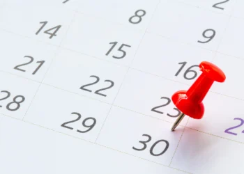 Tacha vermelha fixada no dia 23 do calendário mensal com linhas douradas e números cinzas em fundo branco.