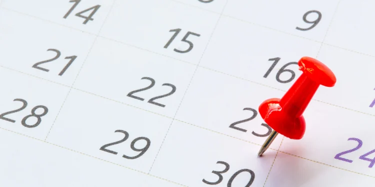 Tacha vermelha fixada no dia 23 do calendário mensal com linhas douradas e números cinzas em fundo branco.