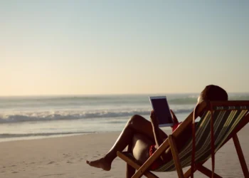 Mulher sentada em cadeira de praia listrada, segurando tablet, observando o mar ao pôr do sol na areia.