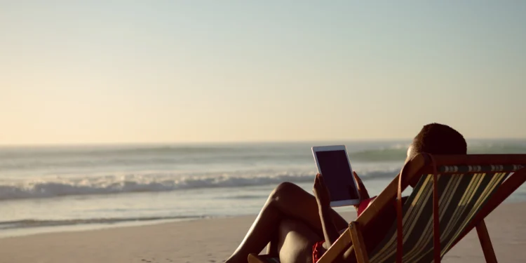 Mulher sentada em cadeira de praia listrada, segurando tablet, observando o mar ao pôr do sol na areia.