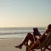 Mulher sentada em cadeira de praia listrada, segurando tablet, observando o mar ao pôr do sol na areia.