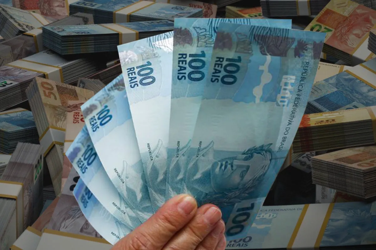 Mão segurando notas de 100 reais com dinheiro ao fundo