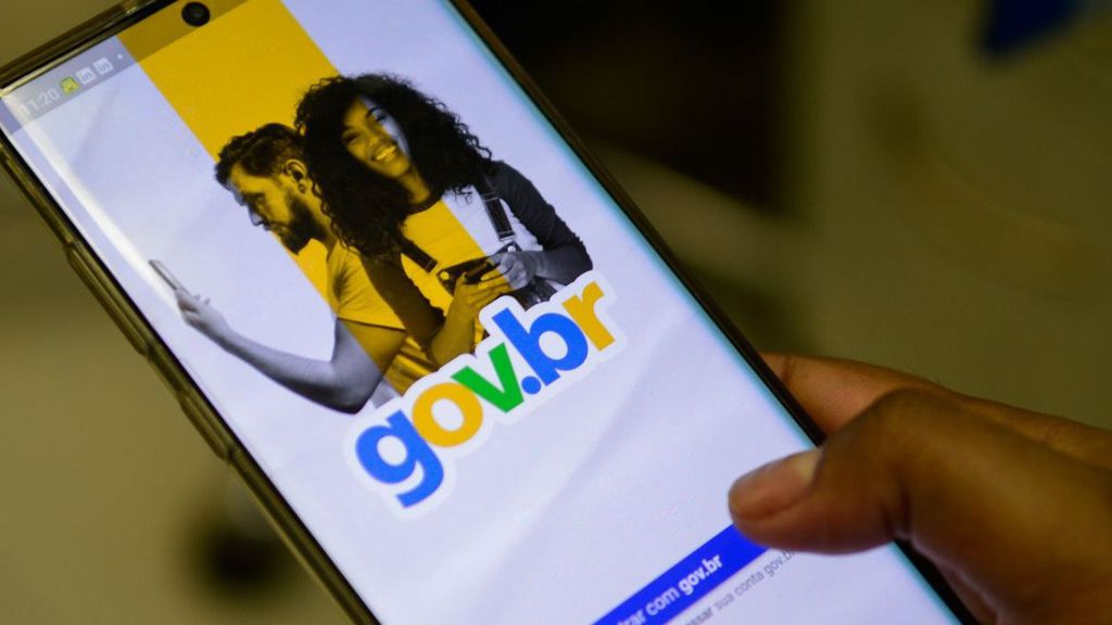 Close de uma mão segurando um smartphone que exibe a tela de login do portal gov.br, com o logotipo colorido da plataforma e a imagem de um casal ao fundo.