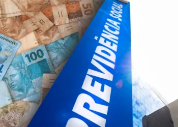 Montagem fotográfica exibindo em primeiro plano uma placa azul vertical com a inscrição "PREVIDÊNCIA SOCIAL" em letras brancas. Ao fundo, diversas cédulas de 50 e 100 reais espalhadas.