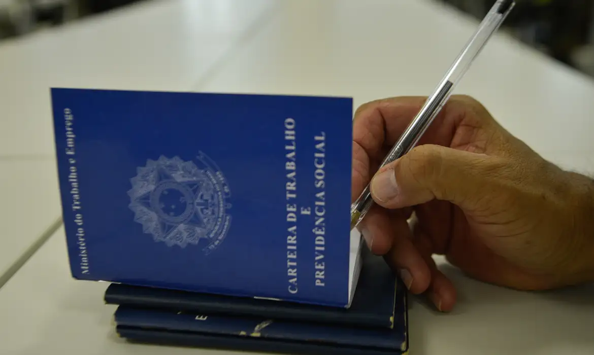 Mão segurando Carteira de Trabalho azul, simbolizando os direitos de pagamento em dobro e folga compensatória para quem trabalha no feriado de 1º de maio