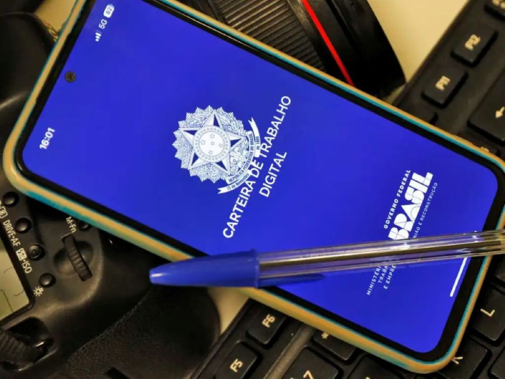 Um smartphone exibindo a tela azul do aplicativo Carteira de Trabalho Digital com o brasão da República. Sobre o celular, repousa uma caneta esferográfica azul, e ao fundo, parte de um teclado de computador e uma câmera fotográfica profissional.