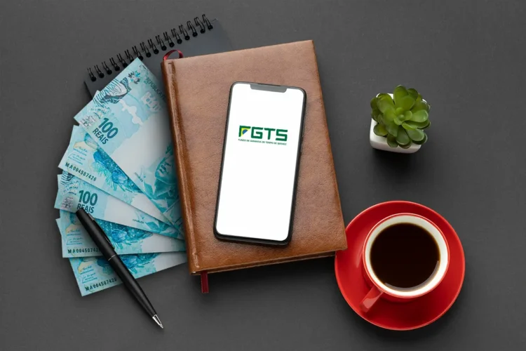 Notas de cem reais, celular com tela do FGTS, caderno marrom, xícara de café e planta pequena sobre mesa escura