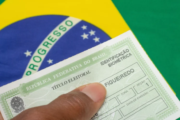 Mão segura título eleitoral da República Federativa do Brasil com marcação de biometria sobre bandeira verde e amarela.