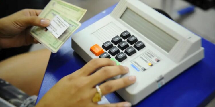 Close das mãos de uma pessoa utilizando uma urna eletrônica de modelo antigo (cor bege). A mão esquerda segura um título de eleitor de papel verde, enquanto o dedo indicador da mão direita pressiona a tecla "Confirma" (verde) no teclado numérico preto.