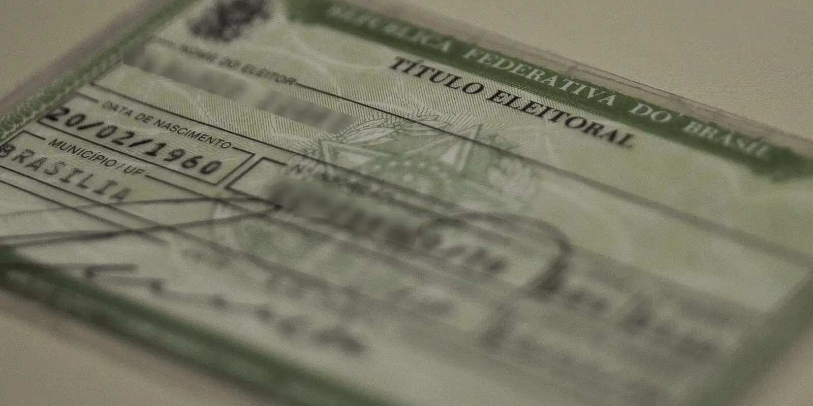 Close detalhado de um Título de Eleitor brasileiro físico, impresso em papel verde e branco. O documento está levemente inclinado e focado, mostrando campos como "Data de Nascimento" (20/02/1960) e "Município/UF" (Brasília). O fundo está desfocado.