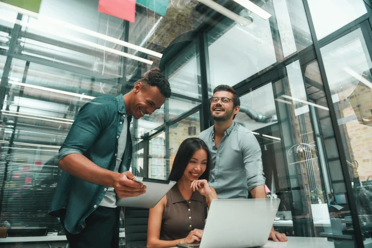 Três profissionais sorriem em escritório com paredes de vidro, tablet e laptop sobre a mesa.