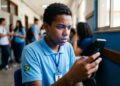 Jovem estudante negro com uniforme azul e bandeira do Brasil verifica celular em corredor escolar com expressão preocupada