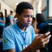 Jovem estudante negro com uniforme azul e bandeira do Brasil verifica celular em corredor escolar com expressão preocupada
