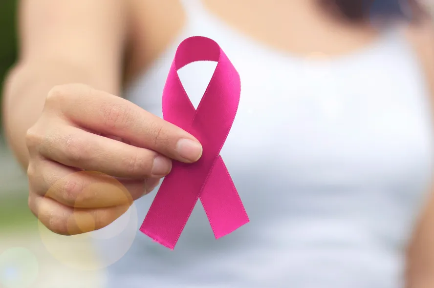 Mulher de regata branca segura laço rosa de cetim em primeiro plano, símbolo da campanha de prevenção ao câncer de mama