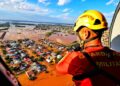 Bombeiro militar com capacete amarelo e colete laranja observa cidade inundada de helicóptero durante operação de resgate