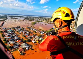 Bombeiro militar com capacete amarelo e colete laranja observa cidade inundada de helicóptero durante operação de resgate