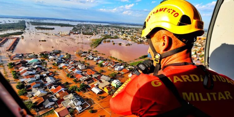 Bombeiro militar com capacete amarelo e colete laranja observa cidade inundada de helicóptero durante operação de resgate