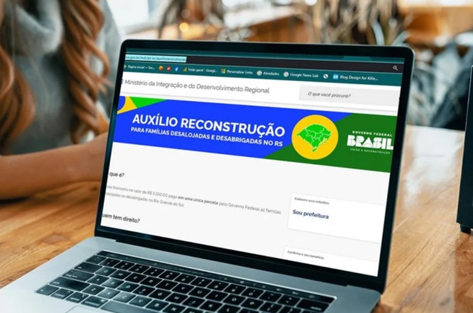 Notebook exibindo site do Auxílio Reconstrução com banner verde e amarelo do Ministério da Integração sobre mesa de madeira