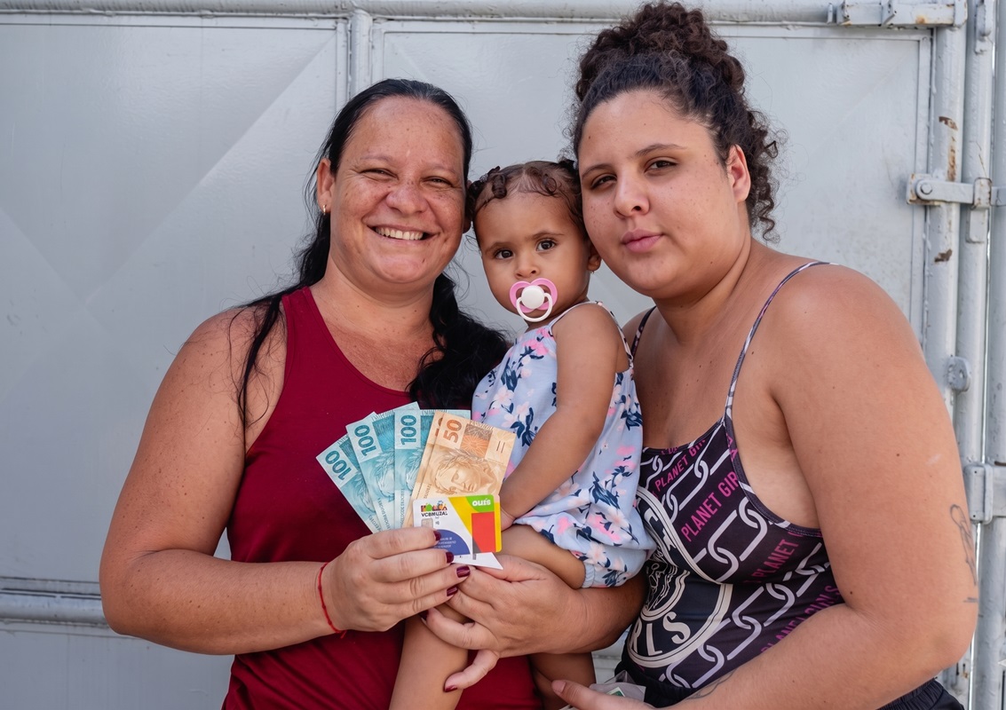 Duas mulheres e um bebê com chupeta mostram cédulas de reais e cartão do Bolsa Família em frente a um portão cinza