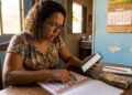 Mulher com cabelos cacheados e óculos consulta celular e calendário de papel, com notas de real sobre a mesa
