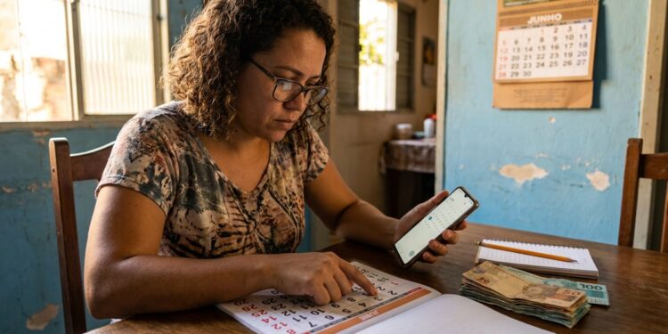 Mulher com cabelos cacheados e óculos consulta celular e calendário de papel, com notas de real sobre a mesa